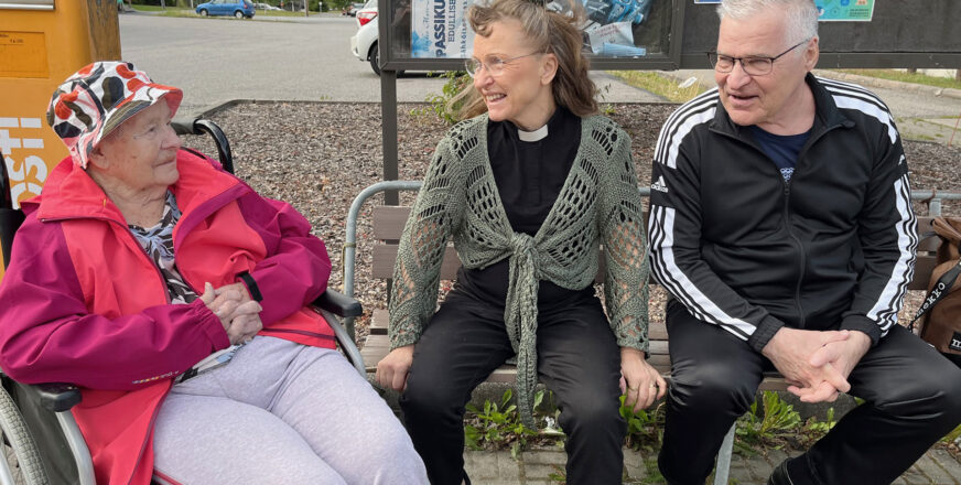 Leena-pappi istuu miehen kanssa torin penkillä. Vasemmalla papin vieressä istuu nainen pyörätuolissa.