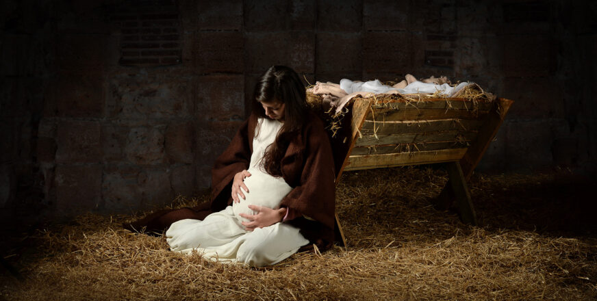Raskaana oleva Neitsyt Maria istuu olkien päällä tallin lattialla, nojaa seimeen ja pitelee vatsaansa.