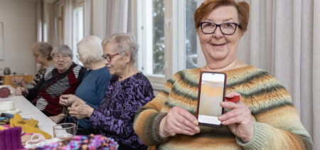 Nainen näyttää kännykästä villapaidan kuvaa. Taustalla muita neulepiiriläisiä neulomassa ja juttelemassa.