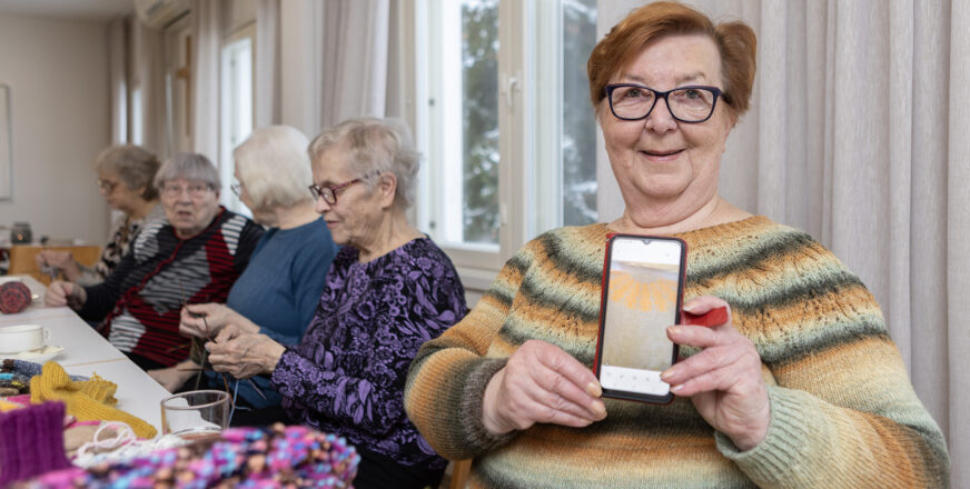 Nainen näyttää kännykästä villapaidan kuvaa. Taustalla muita neulepiiriläisiä neulomassa ja juttelemassa.
