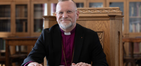 Piispa Jari Jolkkonen istuu hymyillen pöydän ääressä edessään avoinna oleva Raamattu.