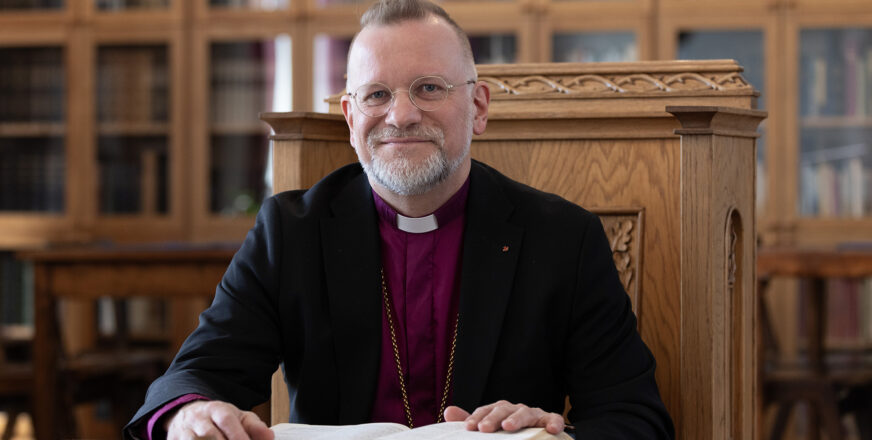 Piispa Jari Jolkkonen istuu hymyillen pöydän ääressä edessään avoinna oleva Raamattu.