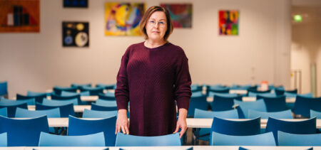 Nainen seisoo koululuokassa pulpettirivien keskellä ja katsoo kameraan.