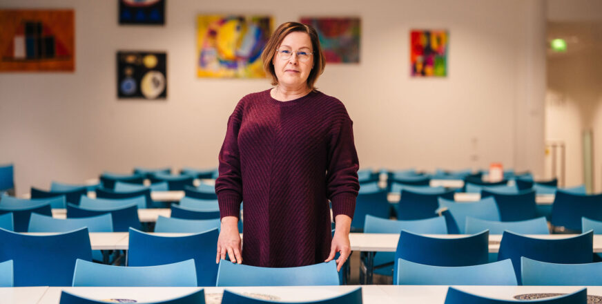Nainen seisoo koululuokassa pulpettirivien keskellä ja katsoo kameraan.