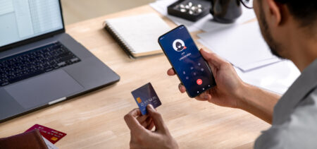 Mies kuvattuna takaapäin, istuu pöydän ääressä. Toisessa kädessä luottokortti ja toisessa kännykkä. Pöydällä myös läppäri.