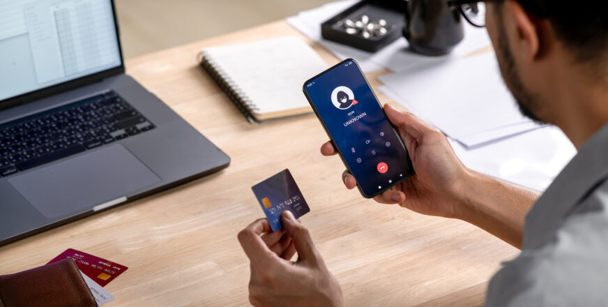 Mies kuvattuna takaapäin, istuu pöydän ääressä. Toisessa kädessä luottokortti ja toisessa kännykkä. Pöydällä myös läppäri.