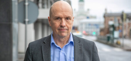 Jyri Komulainen katsoo suoraan kameraan hieman hymyillen, taustalla näkyy harmaata Helsingin katukuvaa.