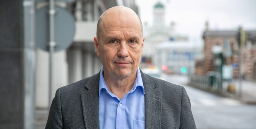 Jyri Komulainen katsoo suoraan kameraan hieman hymyillen, taustalla näkyy harmaata Helsingin katukuvaa.