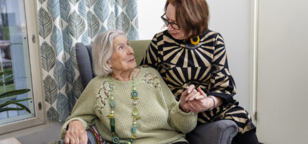 Iäkäs henkilö istuu nojatuolilla ja pitää kädestä nuorempaa henkilöä, joka istuu nojatuolin käsinojalla.