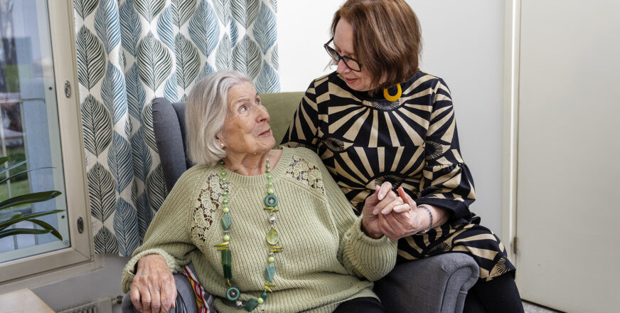 Iäkäs henkilö istuu nojatuolilla ja pitää kädestä nuorempaa henkilöä, joka istuu nojatuolin käsinojalla.