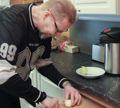 Ihminen pilkkoo sipulia keittiön työtasolla ja hymyilee ruuanlaiton lomassa.