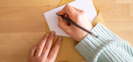 Pöydällä on kirjekuori, josta pilkottaa paperiarkki. Toinen käsi pitää kiinni arkista ja toinen käsi kirjoittaa kirjettä.