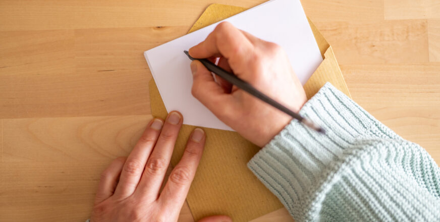 Pöydällä on kirjekuori, josta pilkottaa paperiarkki. Toinen käsi pitää kiinni arkista ja toinen käsi kirjoittaa kirjettä.