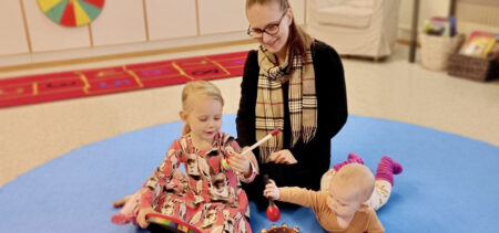 Äiti ja kaksi lasta istuvat matolla soittamassa soittimia.