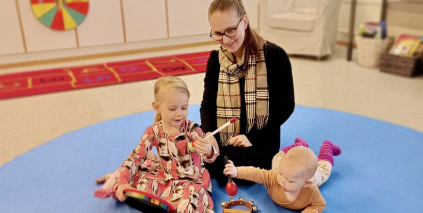 Äiti ja kaksi lasta istuvat matolla soittamassa soittimia.