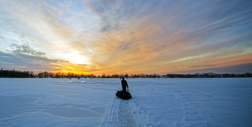 Ihminen kävelee järven jäällä ja vetää varustepulkkaa, taustalla auringonlasku