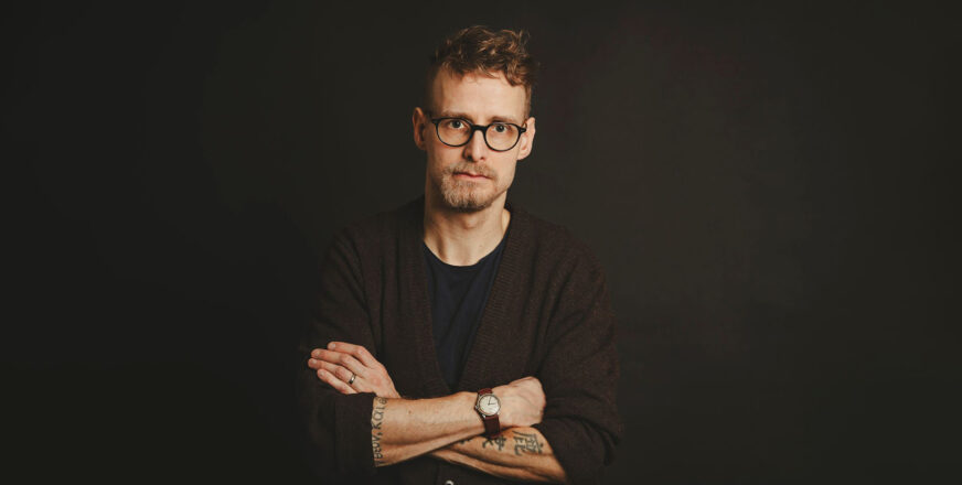 Lari Launonen katsoo kädet puuskassa suoraan kameraan. Hänellä on musta paita ja myös tausta on musta.