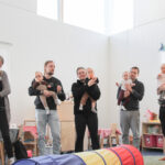 Vanhempia seisoo ja leikkii ringissä lattialla, lapset sylissään.