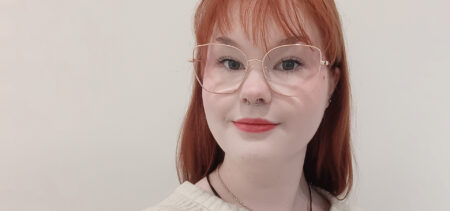 Kasvokuva Emma Riekkisestä, jolla pitkät punaiset hiukset ja silmälasit. Taustalla vaalea seinä.