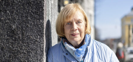 Marketta Rantama nojaa kivimuuriin ja hymyilee hienoisesti, taustalla keväistä ja aurinkoista katukuvaa.