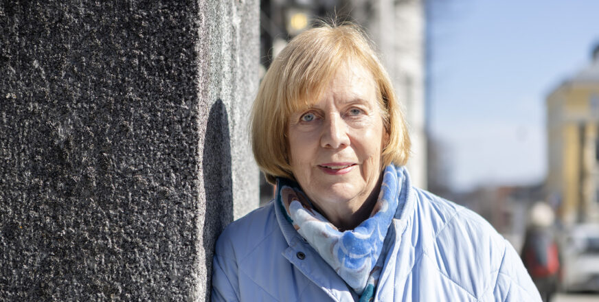 Marketta Rantama nojaa kivimuuriin ja hymyilee hienoisesti, taustalla keväistä ja aurinkoista katukuvaa.