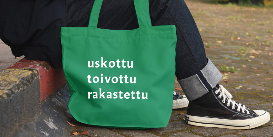 Vihreä kassi, jossa teksti: Uskottu, toivottu, rakastettu. Kassin vierellä istuu ihminen, josta näkyvät vain jalat.