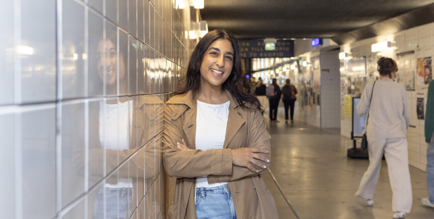 Amani Al-mehsen nojaa asematunnelin seinään ja hymyilee, tunnelissa kulkee ihmisiä.