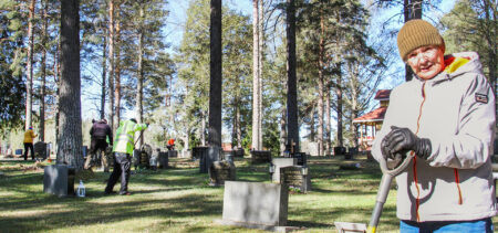 Henkilö nojaa haravaan ja katsoo kameraan, taustalla hautausmaa ja muita vapaaehtoisia.