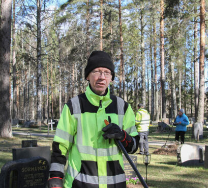 Mies nojaa haravaan työvaatteissaan hautausmaalla.