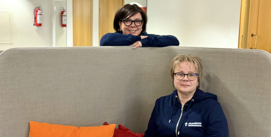 Kiinteistöpalvelupäällikkö Heini Haapalainen istuu sohvalla kirkasväristen tyynyjen kanssa, talousjohtaja Anu Kopponen seisoo sohvan takana nojaten selkänojaan,