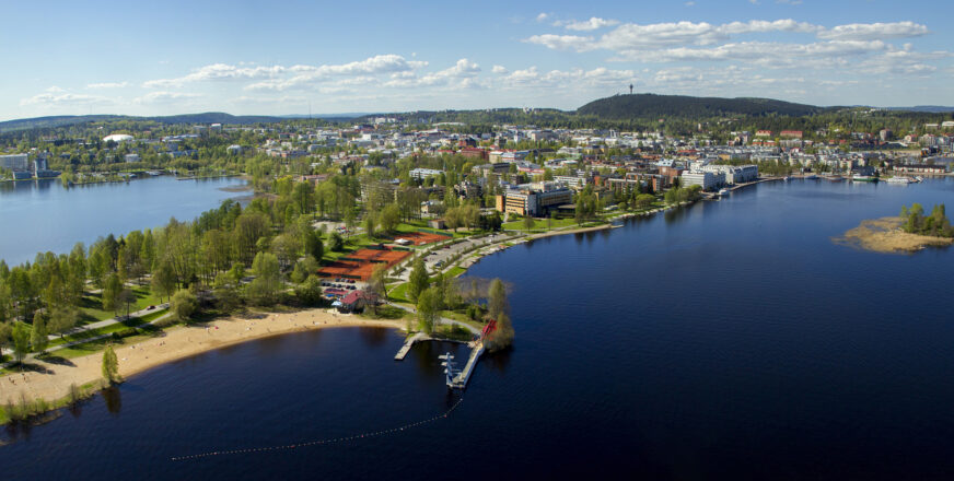 Ilmasta otettu maisemakuva kesäisestä Väinölänniemestä. Ympärillä järveä, taustalla kaupungin keskusta rakennuksineen sekä Puijon mäki ja torni.