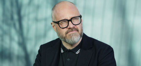 Tuomas Enbuske katsoo mietteliäästi hiukan pois päin kamerasta, taustalla puiden varjoja.
