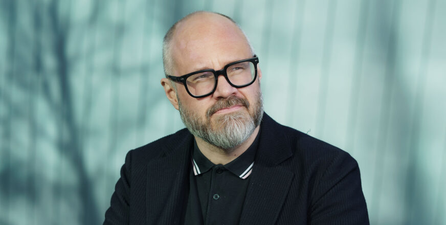Tuomas Enbuske katsoo mietteliäästi hiukan pois päin kamerasta, taustalla puiden varjoja.