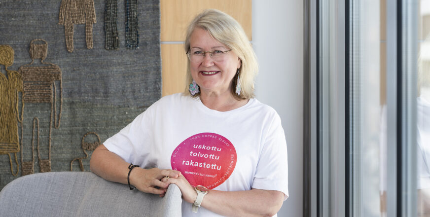 Annmari Salmela seisoo hymyillen nojatuolin takana käsi tuolin selkänojalla.